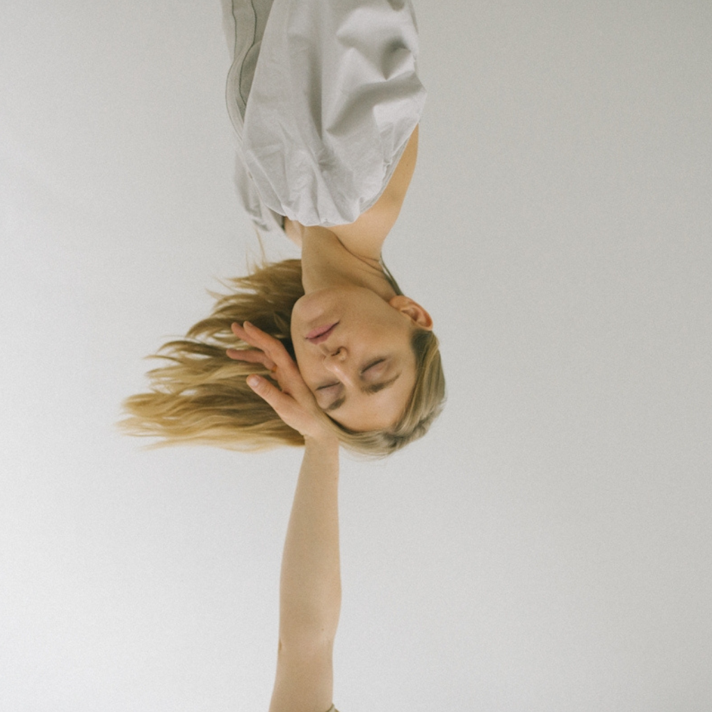 An upside down torso of a woman in white, with blond hair flying to the side. An outstretched arm rests on her cheek from below. She looks peaceful but also upside down.