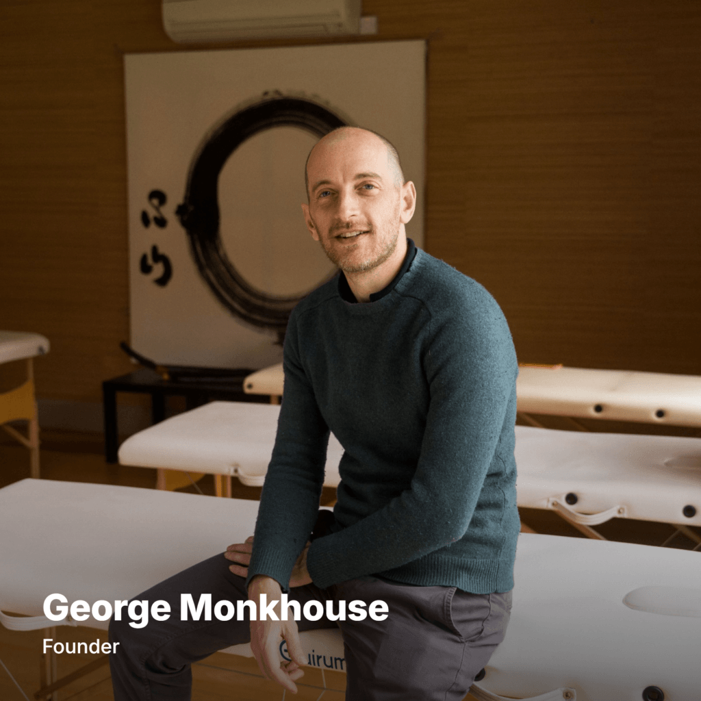George Monkhouse licensed acupuncturist and founder of The Get Well Clinic in a green jumper sitting on a bed in a multibed acupuncture clinic
