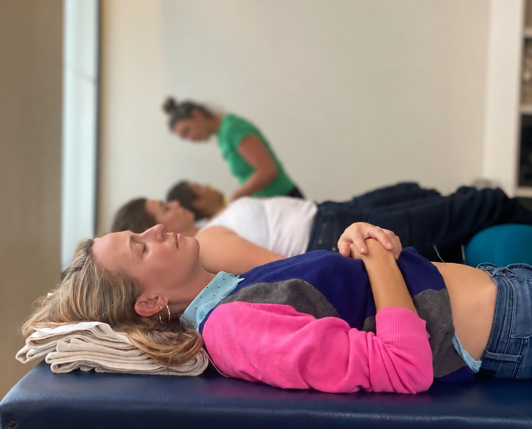 image hero with faces changed a girl in foreground in pink jumper lies on acupuncture table looking rested. in the distance other patients rest and the acupuncturist is standing up at the back treating a patient.