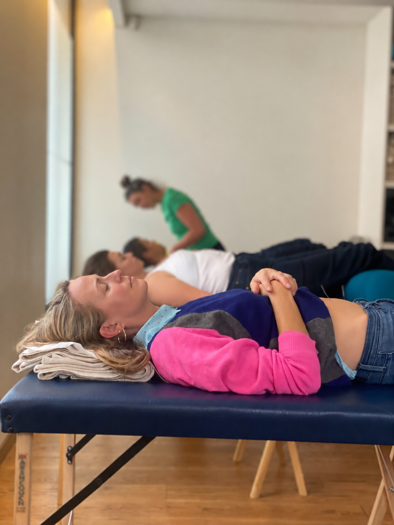 a girl in foreground in pink jumper lies on acupuncture table looking rested. in the distance other patients rest and the acupuncturist is standing up at the back treating a patient.